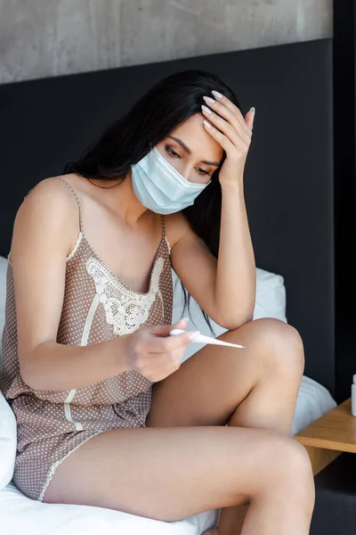 Menina doente em máscara médica olhando para o termômetro enquanto sentado na cama em auto-isolamento — Fotografia de Stock
