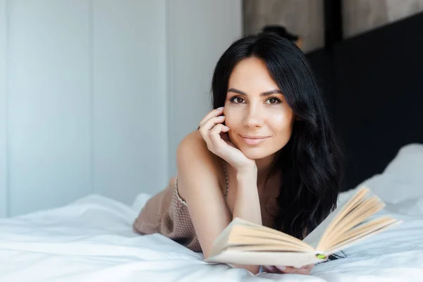 Attractive positive girl reading book on bed at home — Stock Photo