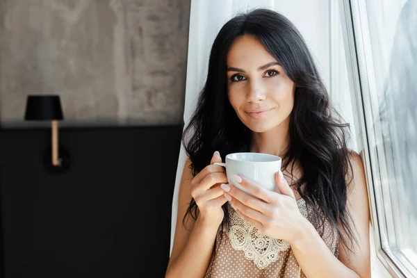 Atractiva mujer joven sosteniendo taza de café cerca de la ventana en casa - foto de stock