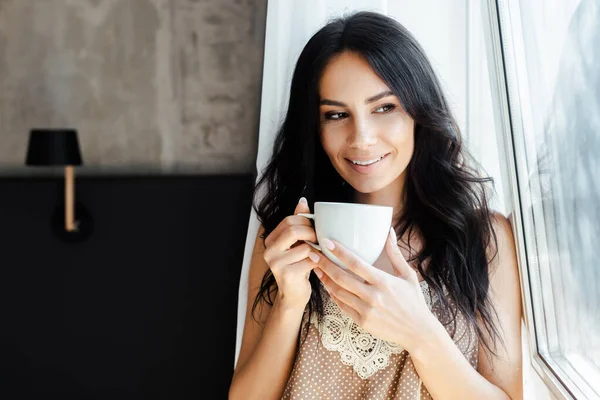 Sonriente joven mujer sosteniendo la taza de café y mirando a través de la ventana - foto de stock