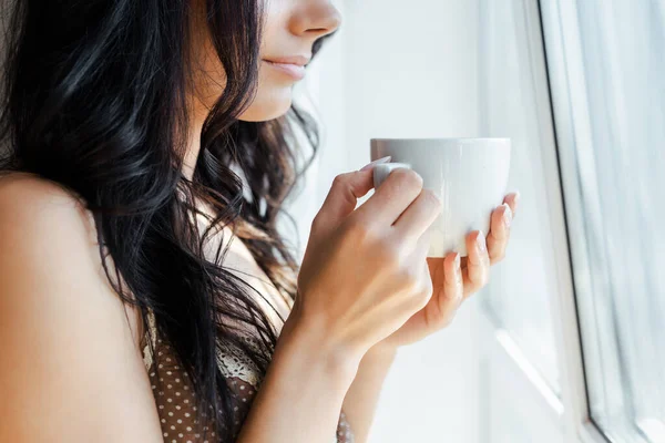 Vista recortada de la niña sosteniendo taza de café cerca de la ventana - foto de stock