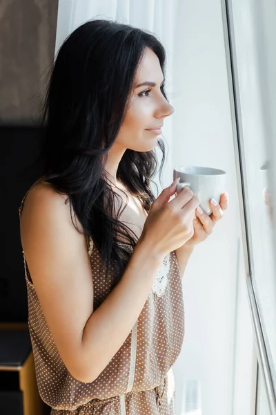 Schönes Mädchen mit Tasse Kaffee und Blick durch das Fenster — Stockfoto