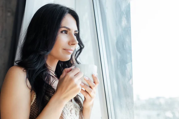 Chica atractiva sosteniendo taza de café y mirando a través de la ventana - foto de stock