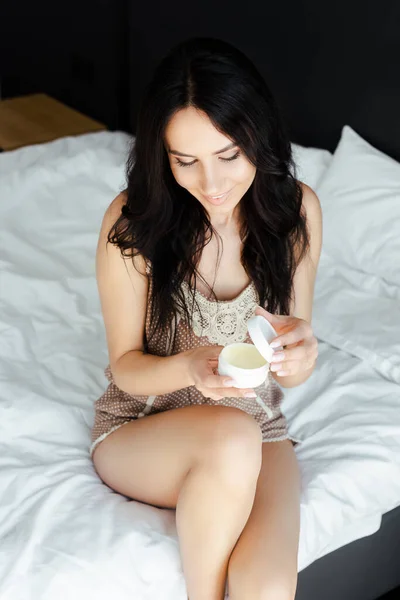 Beautiful smiling woman holding plastic container with cosmetic cream in bedroom — Stock Photo