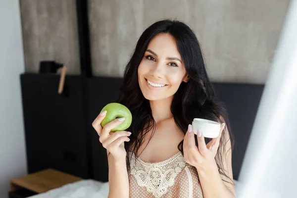 Mulher sorrindo segurando maçã e recipiente de plástico com creme cosmético no quarto — Fotografia de Stock