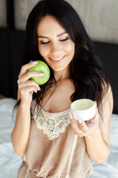 Felice ragazza che tiene mela e contenitore di plastica con crema cosmetica in camera da letto — Foto stock