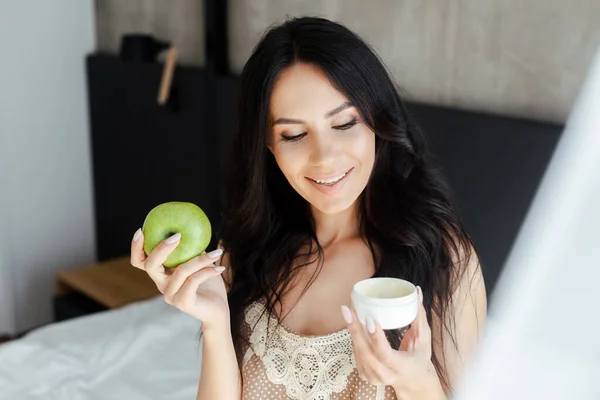 Atractiva mujer sonriente sosteniendo manzana y contenedor de plástico con crema facial en el dormitorio - foto de stock