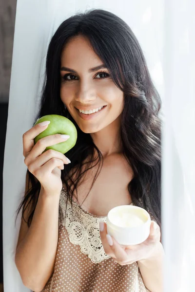 Femme positive tenant pomme verte et récipient en plastique avec crème visage — Photo de stock