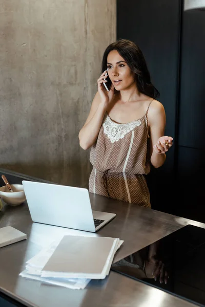 Young woman talking on smartphone and using laptop during breakfast on kitchen with business documents — Stock Photo