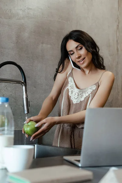 Donna sorridente che parla sullo smartphone e utilizza il computer portatile mentre lava la mela in cucina — Foto stock