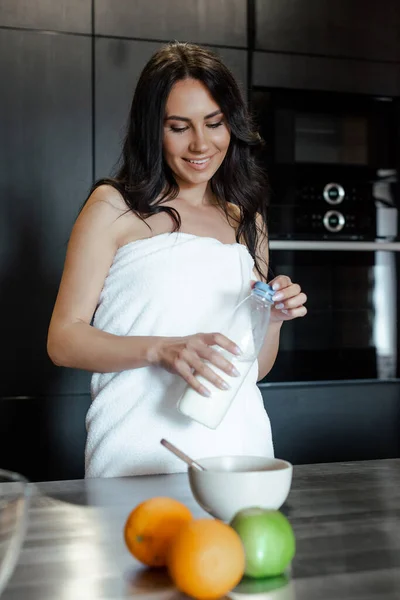 Mujer feliz en toalla haciendo el desayuno con leche en la cocina por la mañana, enfoque selectivo - foto de stock