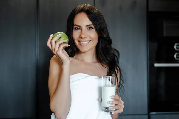 Улыбающаяся женщина в полотенце держит яблоко и стакан норки на кухне утром — стоковое фото