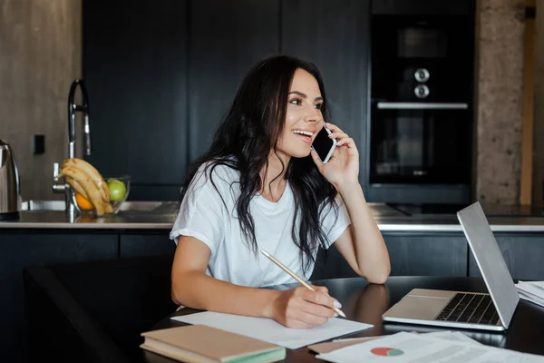 Привлекательная улыбающаяся женщина разговаривает на смартфоне и работает с ноутбуками и деловыми документами на кухне во время самоизоляции — стоковое фото