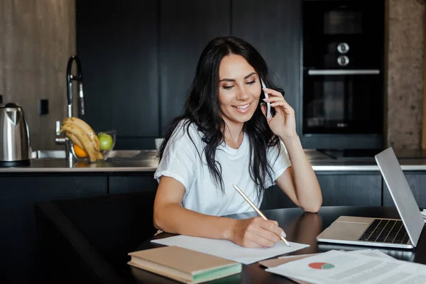 Mujer sonriente hablando en el teléfono inteligente y trabajando con el ordenador portátil y documentos de negocios en la cocina durante el autoaislamiento - foto de stock