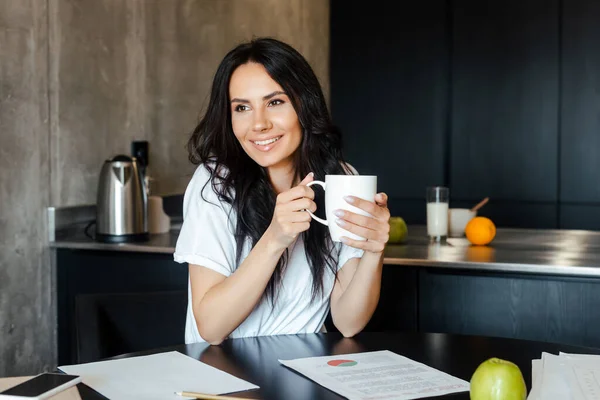 Glückliche Frau mit einer Tasse Kaffee, die zu Hause mit Geschäftsdokumenten über Selbstisolierung arbeitet — Stockfoto