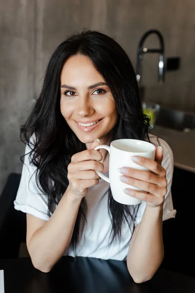 Atractiva chica sonriente sosteniendo taza de café en casa - foto de stock