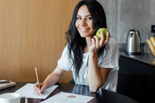 Красивая улыбающаяся женщина работает с деловыми документами и держит яблоко дома на самоизоляции — стоковое фото