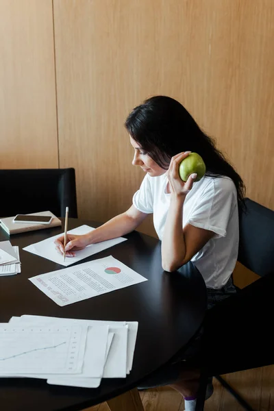 Жінка працює з бізнес-документами і тримає яблуко вдома на самоізоляції — стокове фото