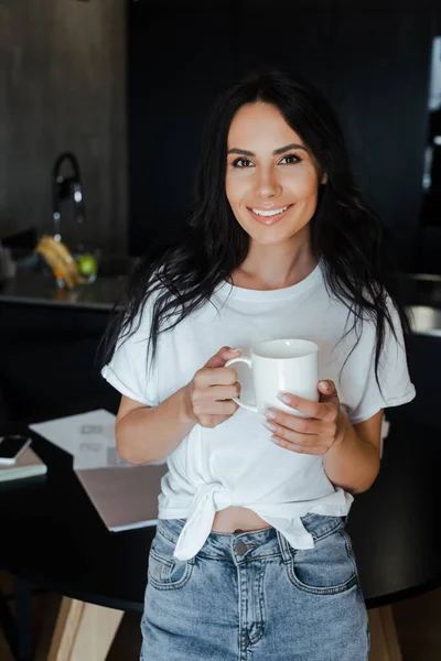 Mujer sonriente sosteniendo una taza de café en casa con documentos de negocios sobre el auto aislamiento - foto de stock