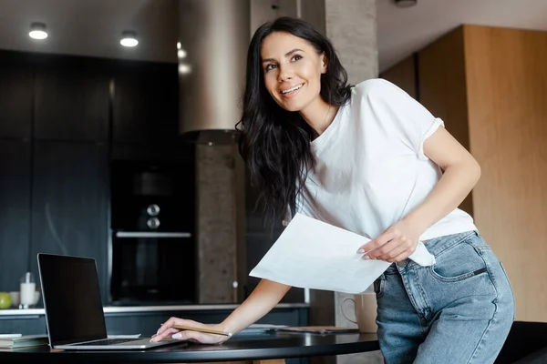 Mujer positiva que trabaja con el ordenador portátil y documentos de negocios en la cocina en el aislamiento personal - foto de stock
