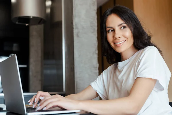 Mulher sorrindo trabalhando com laptop na cozinha em auto-isolamento — Fotografia de Stock