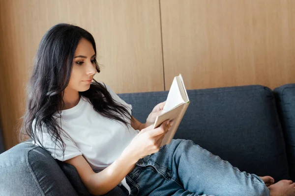 Bella giovane donna che legge libro sul divano a casa — Foto stock