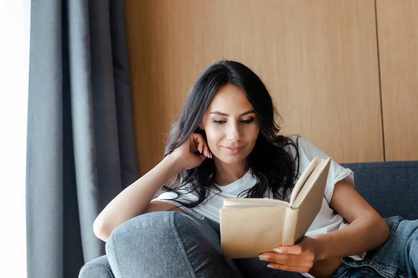 Atractiva chica sonriente leyendo libro en el sofá en casa — Stock Photo