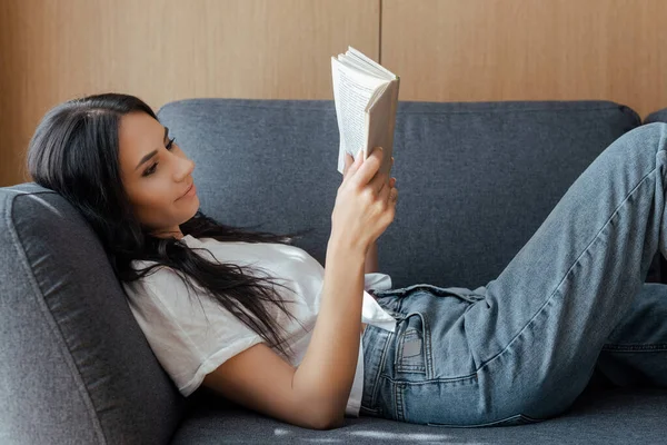 Красивая позитивная девочка, читающая книгу на диване дома — стоковое фото