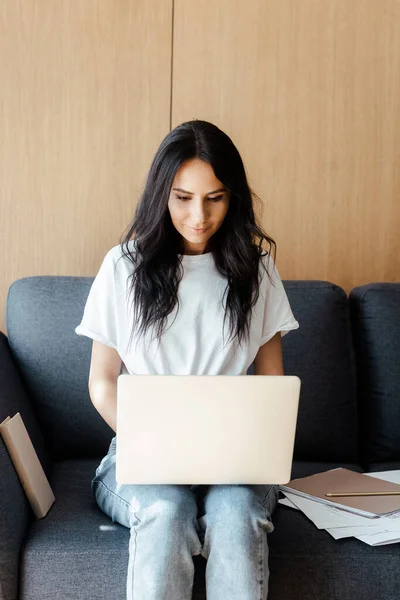 Junge Frau arbeitet während der Selbstisolierung auf dem Sofa am Laptop mit Geschäftsdokumenten — Stockfoto