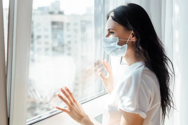 Ill young woman in medical mask looking through window on self isolation — Stock Photo