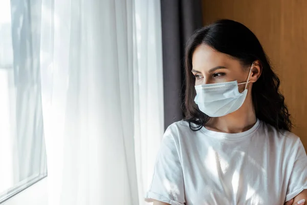 Attraktive kranke Frau in medizinischer Maske, die durch ein Fenster in die Selbstisolierung blickt — Stockfoto