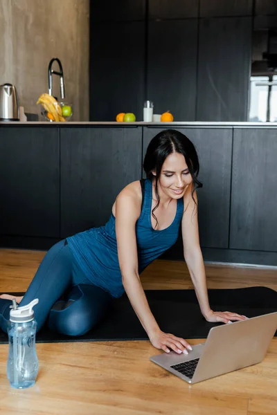 Sorrindo bela esportista sentado no tapete de fitness com garrafa de água e laptop — Fotografia de Stock