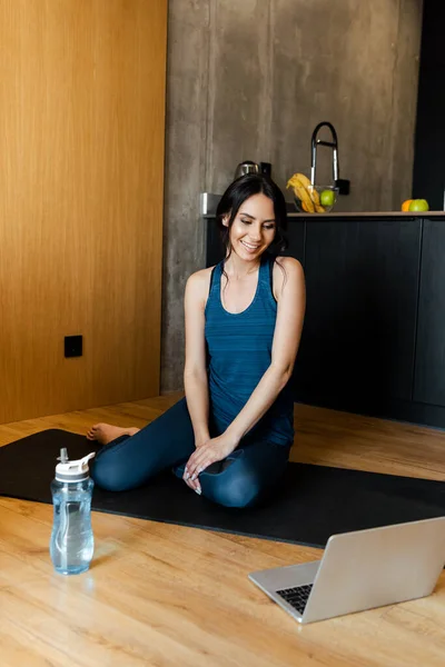 Smiling athletic woman sitting on fitness mat with bottle of water and laptop — Stock Photo