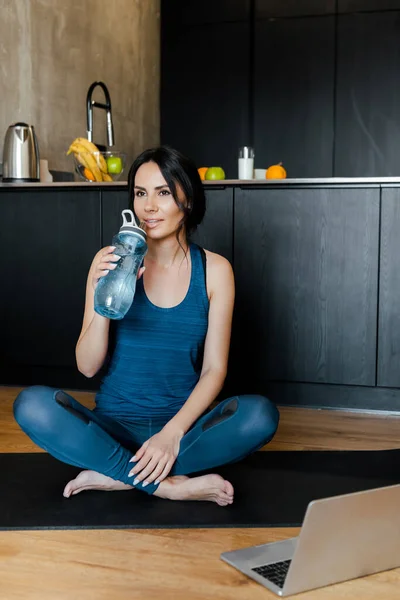 Donna sportiva che beve acqua e siede sul tappetino fitness con laptop — Foto stock