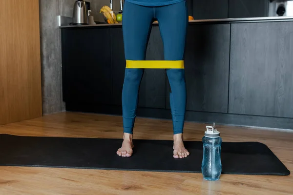 Vista recortada de entrenamiento de mujer atlética con banda de resistencia en alfombra de fitness con botella deportiva - foto de stock