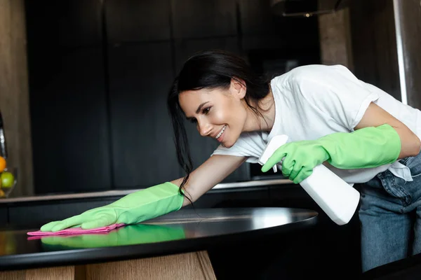 Счастливая привлекательная женщина в латексных перчатках очищающий стол с розовой тряпкой и баллончик — стоковое фото