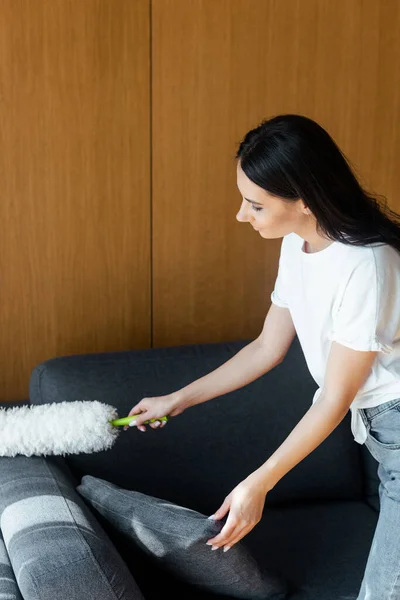 Schöne Frau putzt Staub vom Sofa mit Staubwedel — Stockfoto