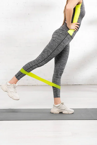 Vista cortada de menina esportiva com a mão no quadril exercício com banda de resistência no tapete de fitness — Fotografia de Stock