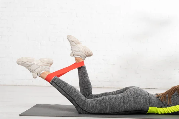 Cropped view of sportive woman lying on fitness mat and working out with resistance band — Stock Photo