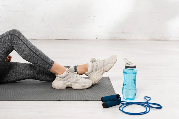 Vista ritagliata della sportiva seduta sul tappeto fitness vicino alla bottiglia sportiva con acqua e corda da salto — Foto stock