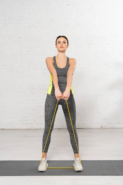 Mujer deportiva mirando a la cámara y haciendo ejercicio con banda de resistencia en la alfombra de fitness - foto de stock