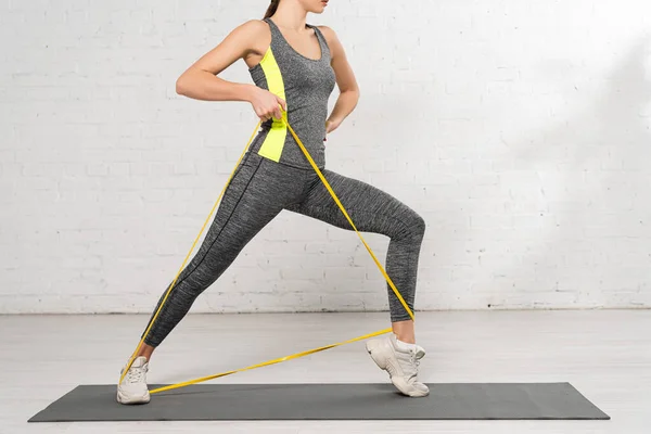 Vista recortada de la mujer deportiva con la mano en la cadera haciendo ejercicio con banda de resistencia en la estera de fitness - foto de stock