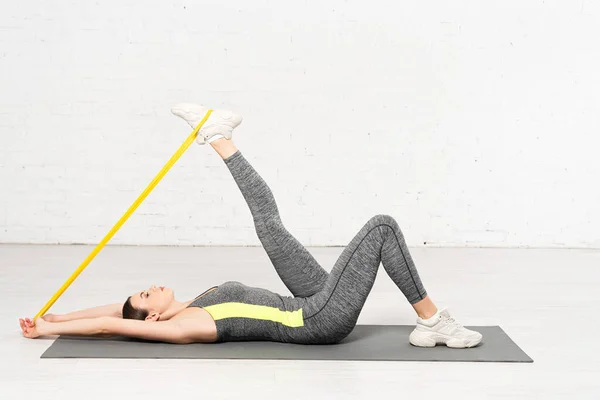 Вид сбоку на женщину в спортивной одежде, выполняющую упражнения с резиновой лентой, лежа на фитнес-коврике у стены из белых бриков — стоковое фото
