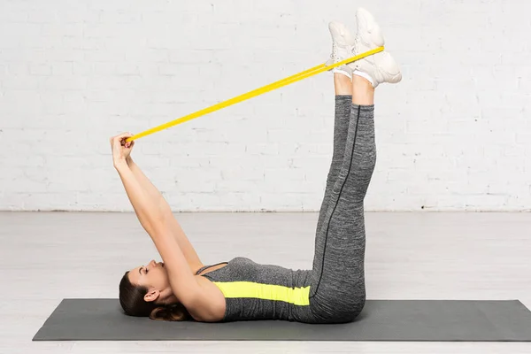 Profilo della donna in abbigliamento sportivo che lavora con la fascia di resistenza mentre giace sul tappeto fitness vicino al muro di mattoni bianchi — Foto stock