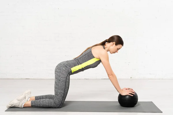 Vue latérale de la fille attrayante en vêtements de sport exercice avec boule noire sur tapis de fitness — Photo de stock