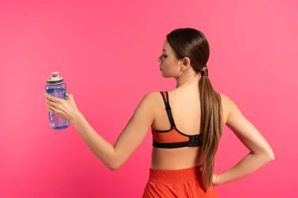 Esportista de pé com a mão no quadril e segurando garrafa de esportes com água isolada em rosa — Fotografia de Stock