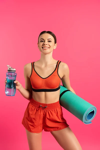 Deportista feliz celebración de la botella de deportes con agua y alfombra de fitness aislado en rosa - foto de stock
