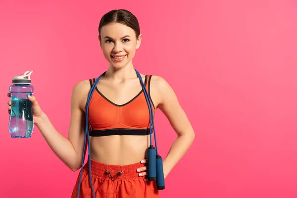 Glückliche Sportlerin hält Sportflasche mit Wasser und steht mit der Hand auf Hüfte isoliert auf rosa — Stockfoto