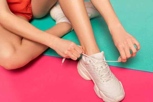 Vista recortada de la mujer sentada en la alfombra de fitness y atando cordones de zapatos en rosa — Stock Photo