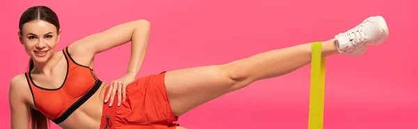 Cultivo panorámico de deportista alegre haciendo ejercicio con banda de resistencia en rosa - foto de stock